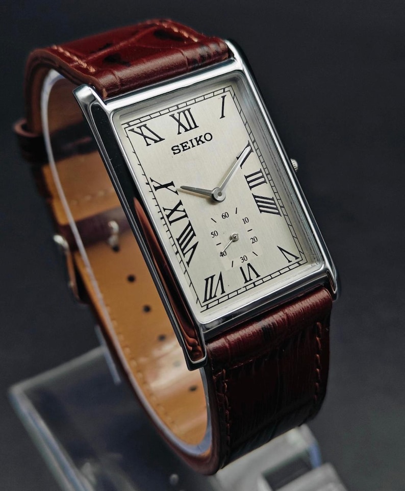 May include: A rectangular Seiko watch with a silver face, black Roman numerals, and a small seconds dial. The watch has a brown leather band. The watch case is silver-toned.