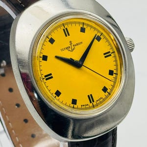 May include: A vintage Ulysse Nardin wristwatch with a bright yellow face and black rectangular hour markers. The watch has a silver-coloured case and a dark brown leather strap. The watch face displays the brand name and an anchor logo.