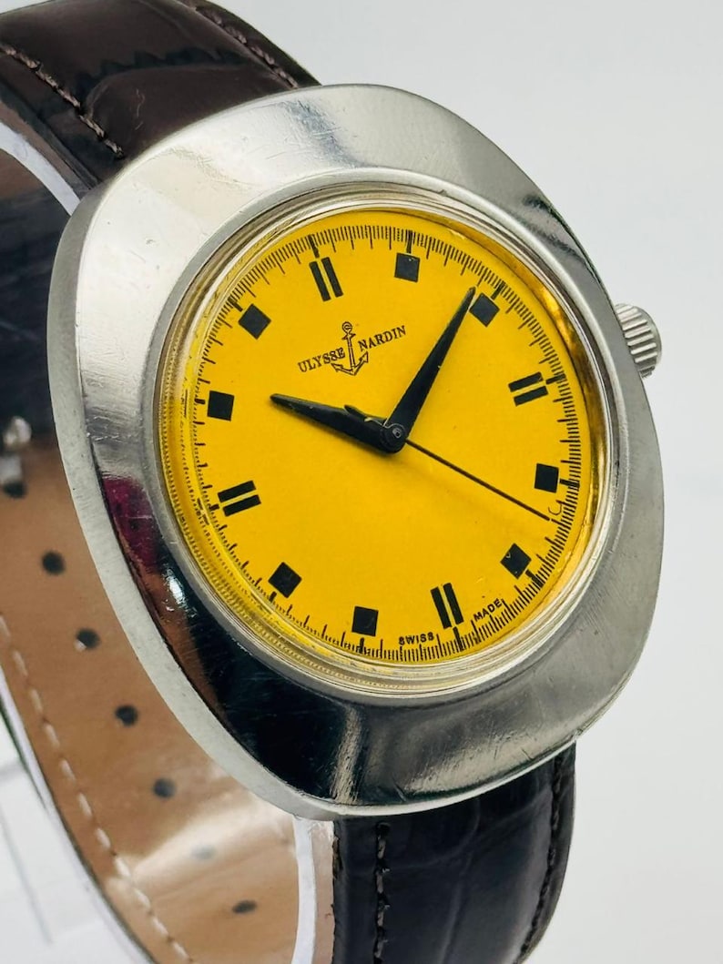 May include: A vintage Ulysse Nardin wristwatch with a bright yellow face and black rectangular hour markers. The watch has a silver-coloured case and a dark brown leather strap. The watch face displays the brand name and an anchor logo.
