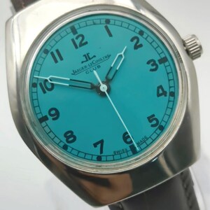 Op de afbeelding: Een zilverkleurig polshorloge met een turquoise wijzerplaat en zwarte cijfers. Het horloge heeft de tekst "JAEGER-LECOULTRE CLUB" en een donkerbruine leren band. De wijzers van het horloge zijn zilver.