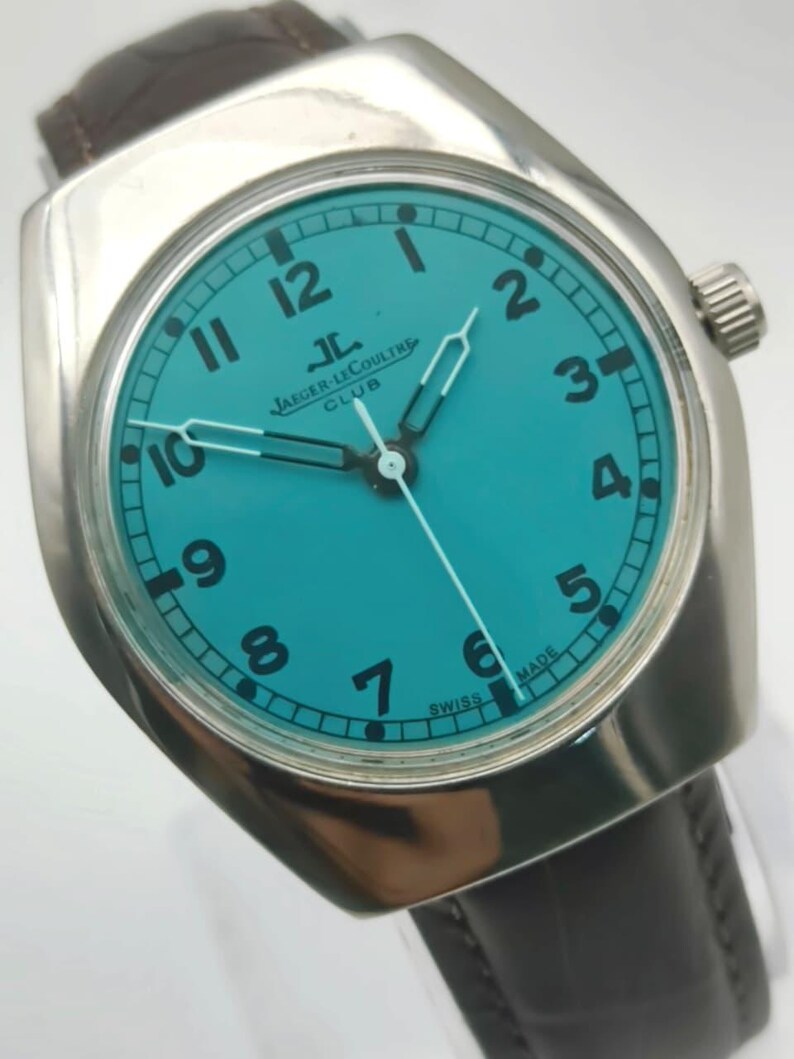 Op de afbeelding: Een zilverkleurig polshorloge met een turquoise wijzerplaat en zwarte cijfers. Het horloge heeft de tekst "JAEGER-LECOULTRE CLUB" en een donkerbruine leren band. De wijzers van het horloge zijn zilver.