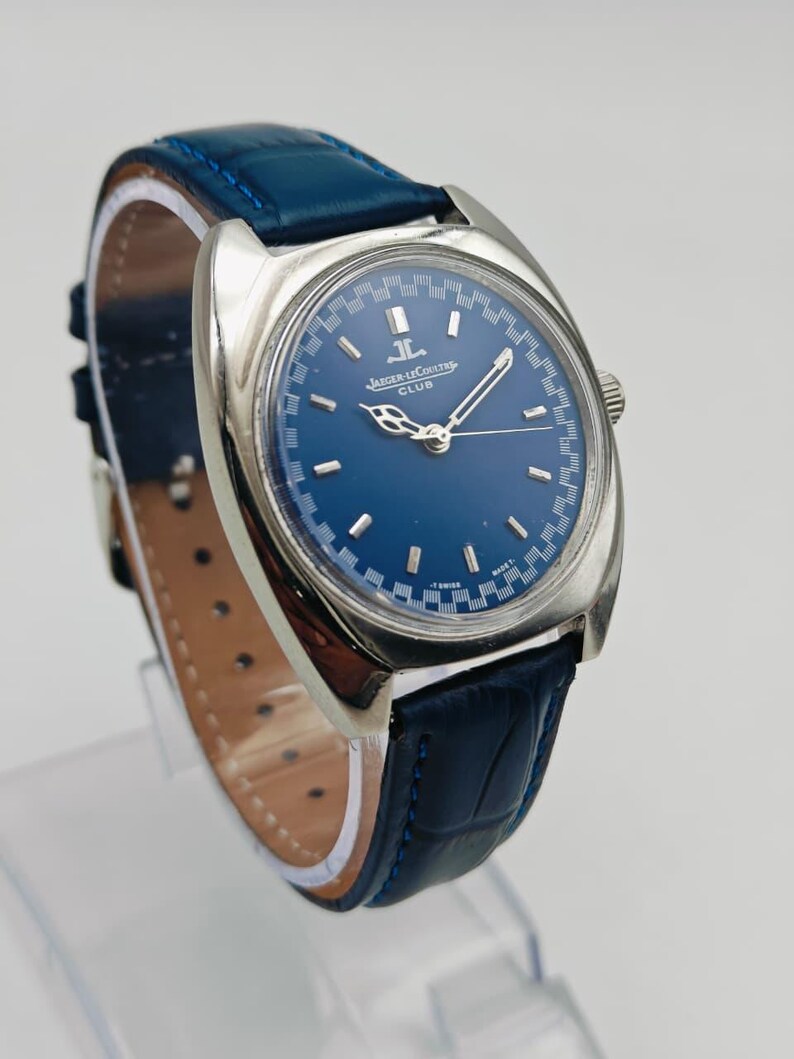 Puede incluir: Un reloj de pulsera plateado con esfera azul y correa de cuero azul. La esfera del reloj presenta el texto "JAEGER-LECOULTRE CLUB". La correa mide aproximadamente 2 cm de ancho. El reloj se muestra sobre un soporte transparente.