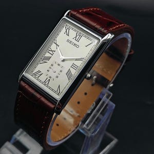 May include: A rectangular Seiko wristwatch with a silver case and a cream-colored face featuring Roman numerals. The watch has a brown leather band. The watch is displayed on a clear stand against a dark background.