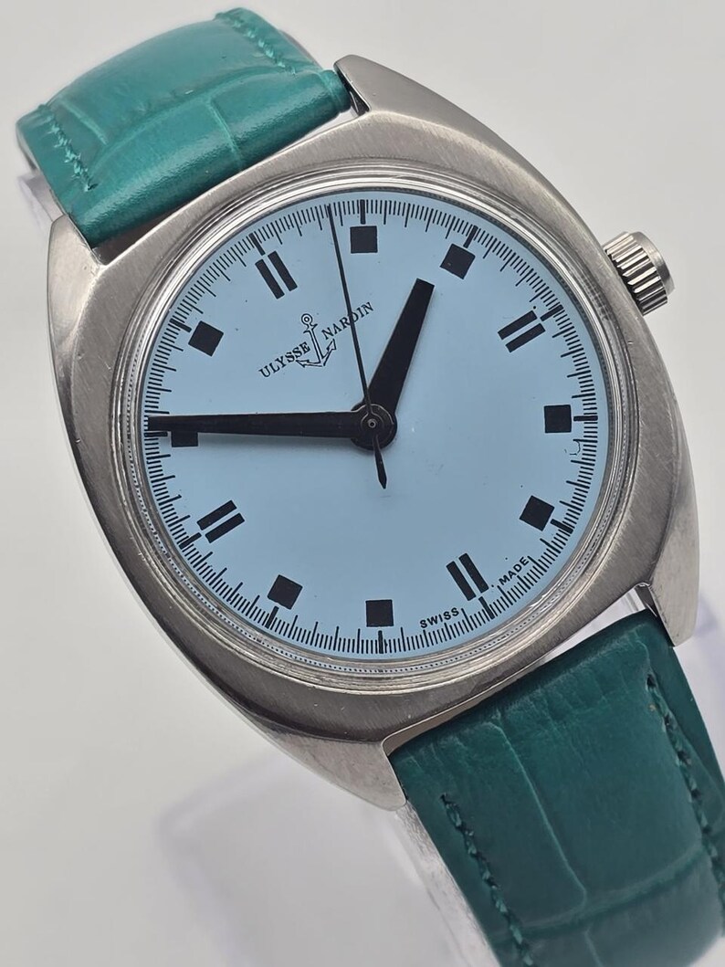 Peut inclure: Une montre Ulysse Nardin vintage avec un cadran bleu clair et des aiguilles et marqueurs noirs. La montre a un bo&icirc;tier argent&eacute; et un bracelet en cuir vert d'eau. Le cadran affiche le logo Ulysse Nardin et l'inscription "SWISS MADE".