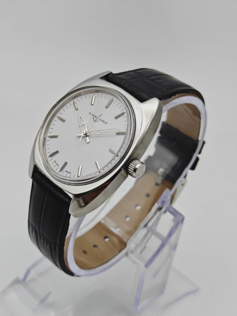 May include: A silver-toned wristwatch with a white face and black leather band. The watch features a round face with silver hands and markers. The band has a crocodile pattern. The watch is displayed on a clear stand.
