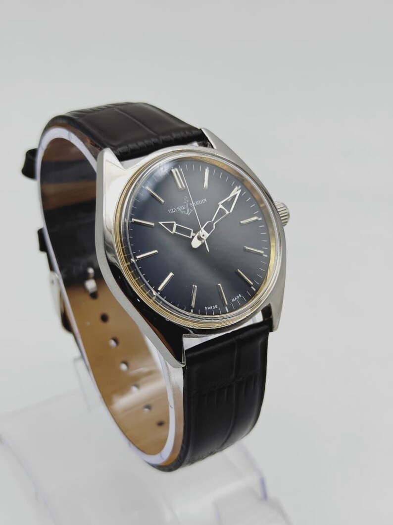 May include: A vintage Ulysse Nardin wristwatch with a dark blue face and silver hands. The watch has a silver case and a black leather band. The watch is displayed on a clear stand.