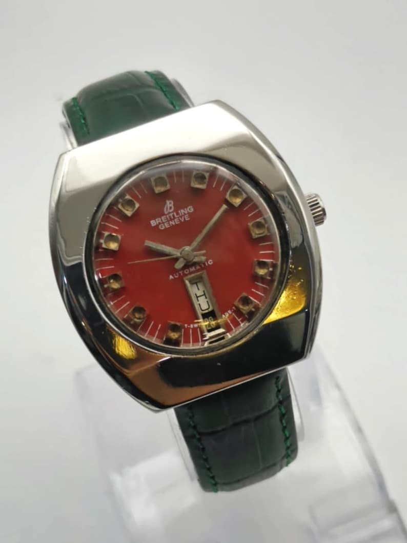 May include: A vintage Breitling Geneve automatic watch with a red dial and a date window. The watch has a silver-tone case and a green leather strap. The watch face features gold-tone hour markers and hands.