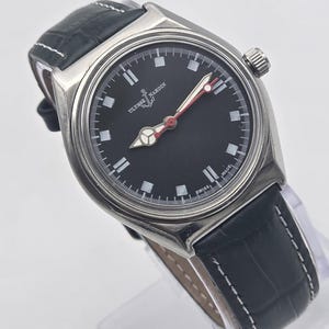 Puede incluir: Un reloj Ulysse Nardin de acero inoxidable con esfera negra, marcadores blancos y agujas con punta roja. El reloj tiene una correa de cuero negro con costuras blancas. La esfera del reloj dice "Ulysse Nardin" y "Swiss Made".