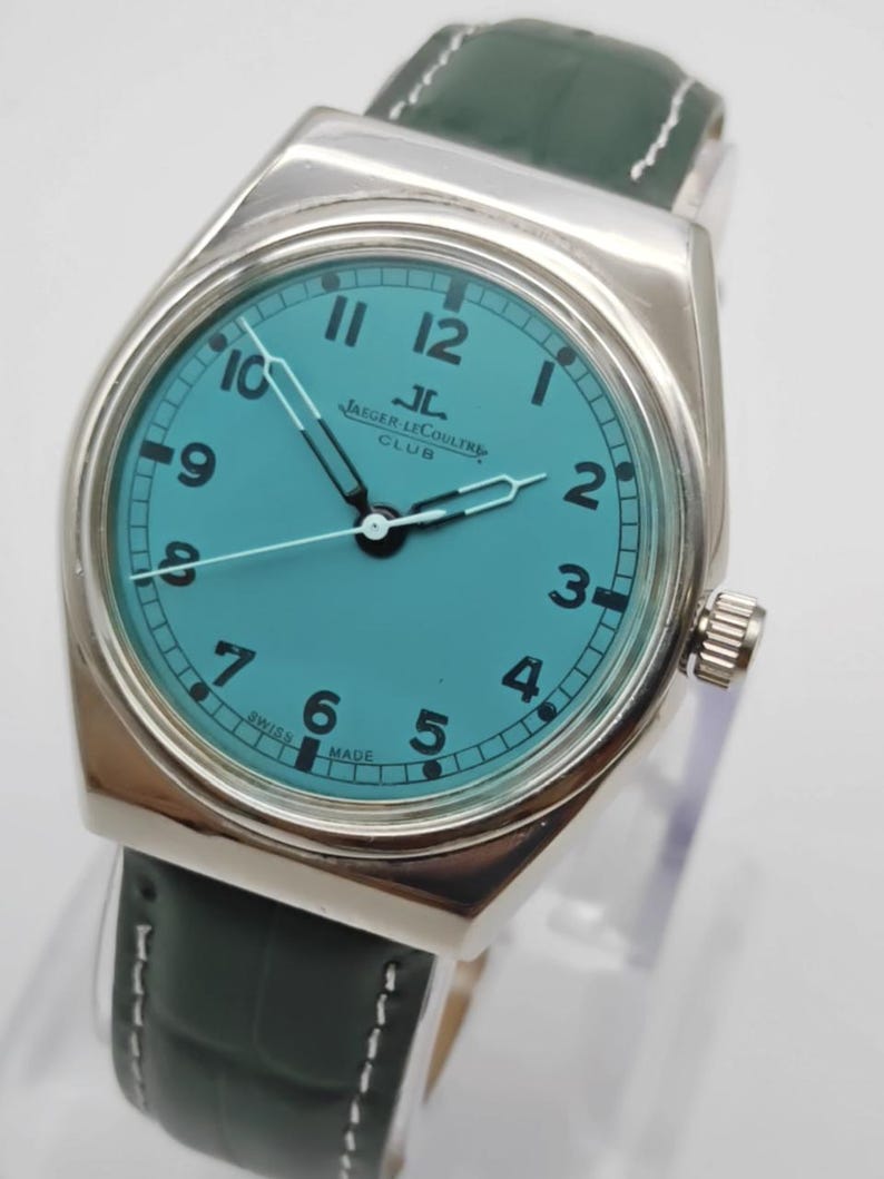 Pode incluir: Um rel&oacute;gio prateado com mostrador turquesa e algarismos pretos. O rel&oacute;gio apresenta o texto "JAEGER LECOULTRE CLUB" e tem uma bracelete em pele verde escura. Os ponteiros do rel&oacute;gio s&atilde;o prateados. O rel&oacute;gio &eacute; um rel&oacute;gio cl&aacute;ssico.