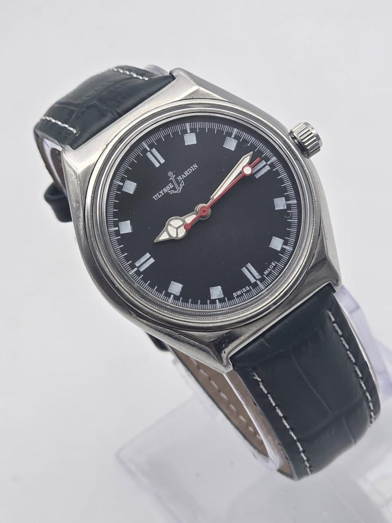 Puede incluir: Un reloj Ulysse Nardin de acero inoxidable con esfera negra, marcadores blancos y manecillas con punta roja. El reloj tiene una correa de cuero negro con costuras blancas. La esfera del reloj dice "Ulysse Nardin" y "Swiss Made".