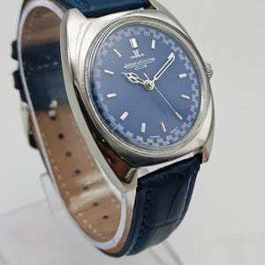 Puede incluir: Un reloj Jaeger-LeCoultre Club plateado con esfera azul y correa de cuero azul. La esfera del reloj presenta agujas e indicadores plateados. La correa mide aproximadamente 2 cm de ancho.