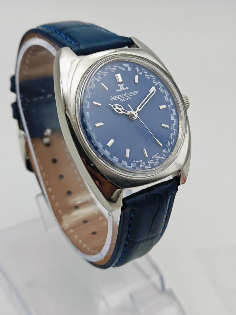 Puede incluir: Un reloj Jaeger-LeCoultre Club plateado con esfera azul y correa de cuero azul. La esfera del reloj presenta agujas e indicadores plateados. La correa mide aproximadamente 2 cm de ancho.