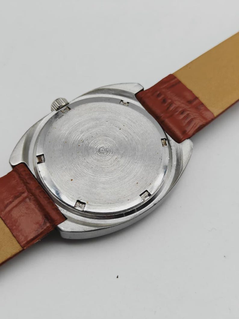 May include: A vintage wristwatch with a silver-colored case and a brown leather strap. The watch face is not visible. The back of the watch has a circular pattern. The strap is 0.75 inches wide.