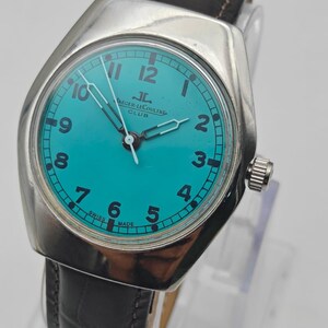 Op de afbeelding: Een vintage Jaeger-LeCoultre Club horloge met een turquoise wijzerplaat en zilveren kast. Het horloge heeft zwarte cijfers, zilveren wijzers en een bruine leren band. De wijzerplaat leest "Jaeger-LeCoultre Club" en "Swiss Made".