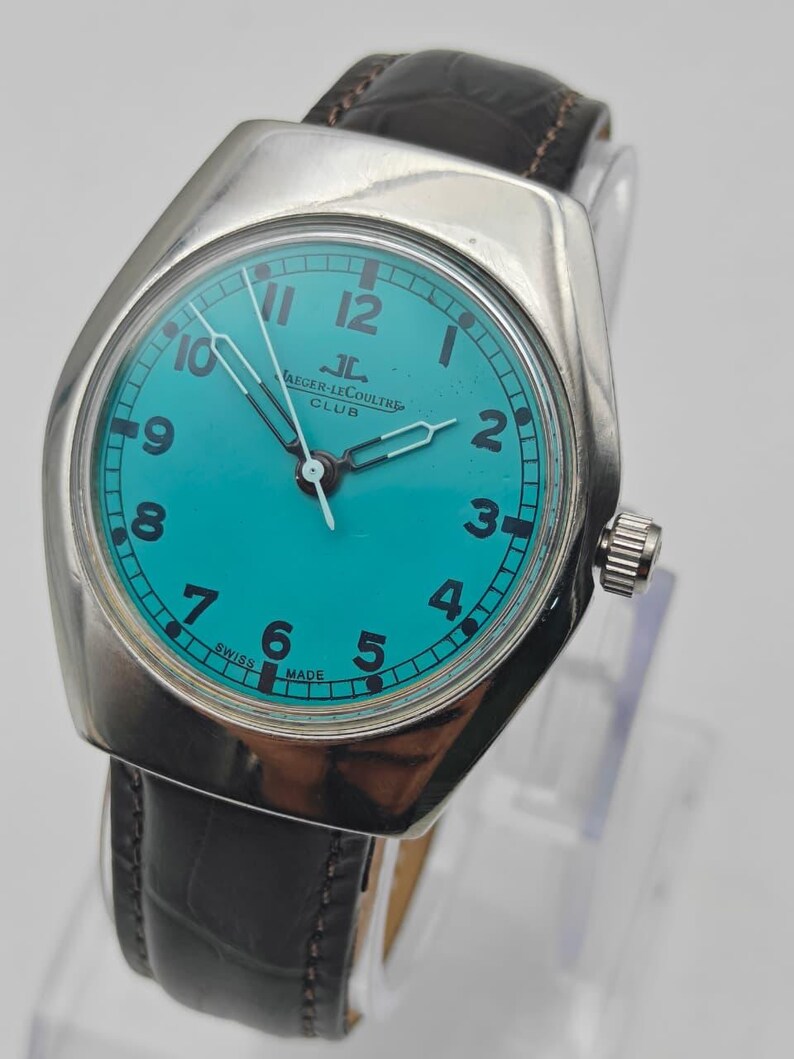 Op de afbeelding: Een vintage Jaeger-LeCoultre Club horloge met een turquoise wijzerplaat en zilveren kast. Het horloge heeft zwarte cijfers, zilveren wijzers en een bruine leren band. De wijzerplaat leest "Jaeger-LeCoultre Club" en "Swiss Made".
