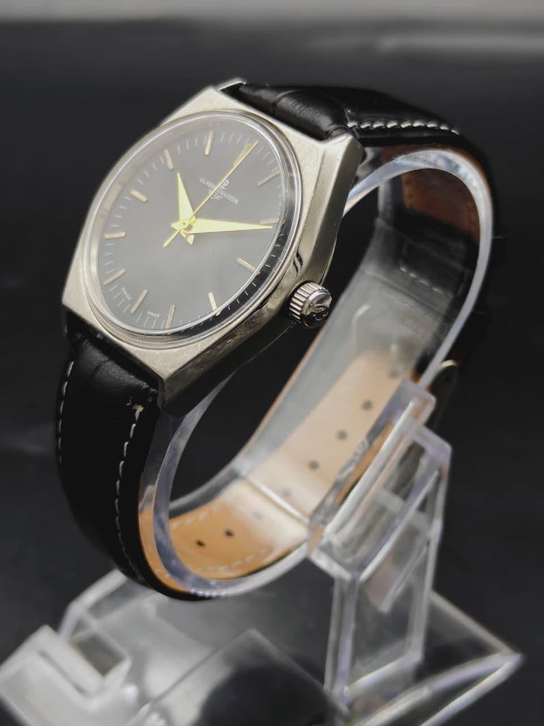 May include: A wristwatch with a silver-toned case and a round black dial featuring gold-colored hands and hour markers. The watch is attached to a black leather strap with white stitching and a silver crown on the side of the case.