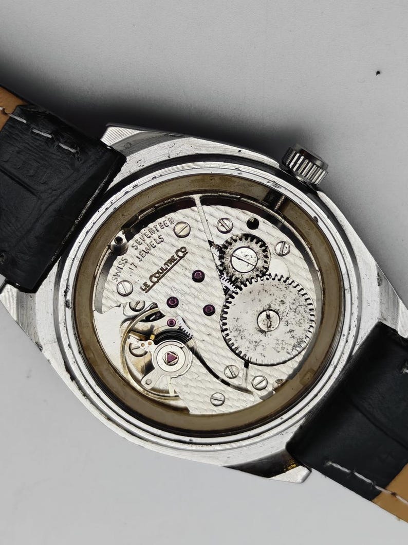 Pode incluir: Vista detalhada do mecanismo de um rel&oacute;gio de pulso vintage. A caixa prateada revela a intrincada maquinaria, com uma bracelete de couro preta. O texto diz "SWISS SEVENTEEN JEWELS LE COULTRE CO."