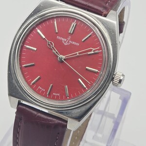 May include: A vintage Ulysse Nardin wristwatch with a rectangular silver case and a vibrant red dial. The watch features silver hands and markers, a burgundy leather strap, and the text "SWISS MADE".