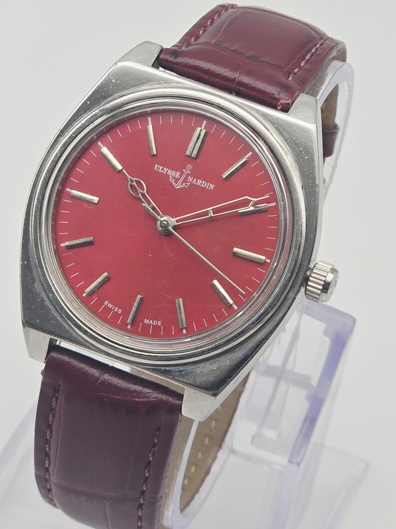 May include: A vintage Ulysse Nardin wristwatch with a rectangular silver case and a vibrant red dial. The watch features silver hands and markers, a burgundy leather strap, and the text "SWISS MADE".