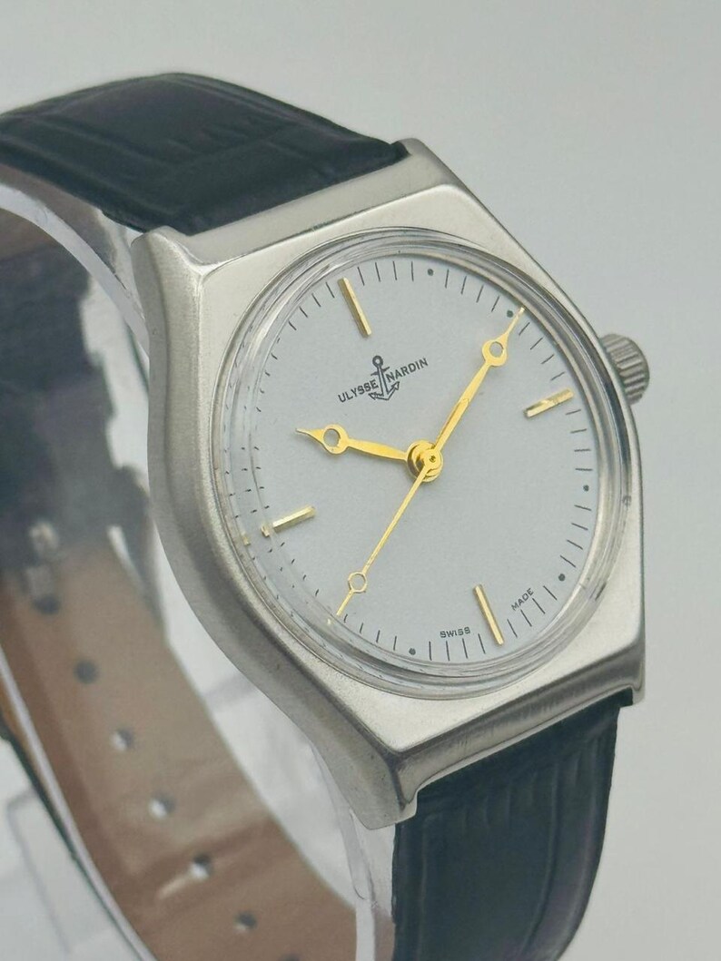 May include: A silver-toned Ulysse Nardin wristwatch with a light gray face, gold hands, and a black leather band. The watch face features the Ulysse Nardin logo and the word "SWISS". The watch has a rectangular shape.