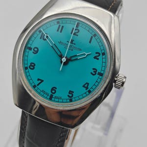 Op de afbeelding: Een zilverkleurig Jaeger-LeCoultre Club horloge met een turquoise wijzerplaat en zwarte wijzers. Het horloge heeft een zwarte leren band en een zilveren kroon. De wijzerplaat toont de cijfers 1 tot en met 12. De woorden "SWISS MADE" staan op de wijzerplaat.