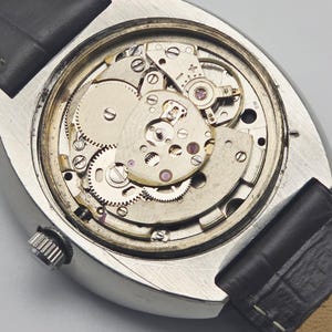 Puede incluir: Primer plano del mecanismo expuesto de un reloj de pulsera vintage. El reloj presenta una caja plateada, una correa de cuero negro y engranajes y mecanismos intrincados. Los componentes internos del reloj son visibles.