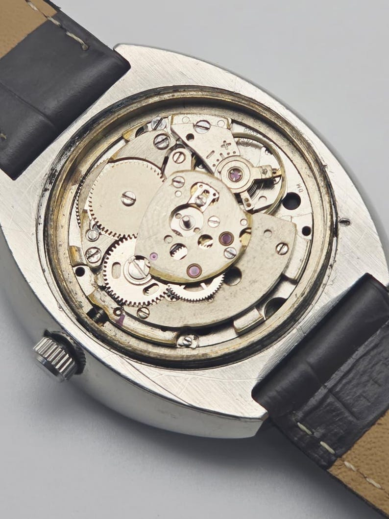 Puede incluir: Primer plano del mecanismo expuesto de un reloj de pulsera vintage. El reloj presenta una caja plateada, una correa de cuero negro y engranajes y mecanismos intrincados. Los componentes internos del reloj son visibles.