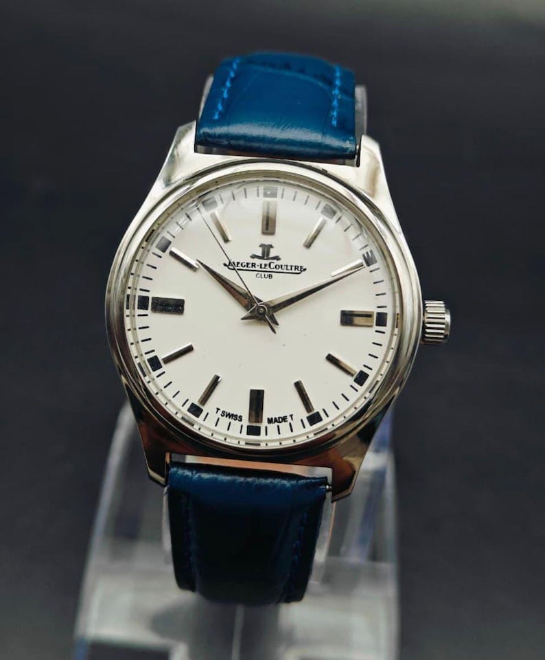 Peut inclure: Une montre Jaeger-LeCoultre Club argent&eacute;e avec un cadran blanc et un bracelet en cuir bleu. Le cadran comporte des marqueurs et des aiguilles noirs, avec le nom de la marque et le texte "T SWISS MADE T". Le bracelet est bleu vif.