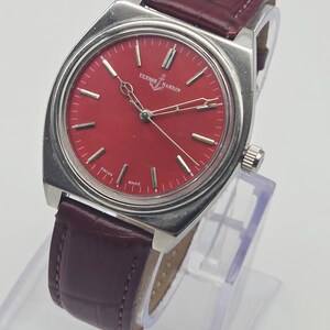 May include: A vintage Ulysse Nardin wristwatch with a red dial and silver-tone case. The watch features silver hands and markers, and a burgundy leather strap. The watch is displayed on a clear stand. The dial reads "Ulysse Nardin" and "Swiss Made".