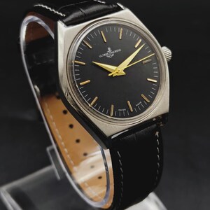 May include: A wristwatch with a silver-toned case, a black dial, and gold-colored hands and hour markers. The dial features the text Ulysse Nardin and Swiss Made. It is fitted with a black leather strap with white stitching and a light-colored lining.