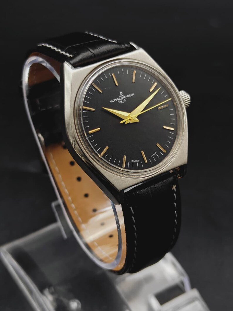 May include: A wristwatch with a silver-toned case, a black dial, and gold-colored hands and hour markers. The dial features the text Ulysse Nardin and Swiss Made. It is fitted with a black leather strap with white stitching and a light-colored lining.