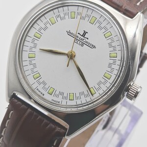 May include: A silver-toned Jaeger-LeCoultre Club watch with a white face and gold hands. The watch has a brown leather band. The face displays the brand name and the words "Swiss Made".
