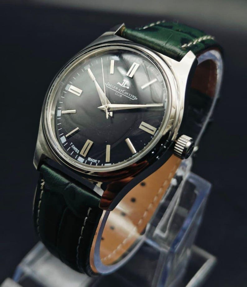 K&ouml;nnte beinhalten: Eine silberfarbene Armbanduhr mit einem dunkelgrauen Zifferblatt und einem gr&uuml;nen Lederarmband. Das Zifferblatt hat silberne Zeiger und Markierungen, mit dem Aufdruck "JAEGER-LECOULTRE CLUB". Das Armband ist aus gr&uuml;nem Leder mit Krokodilmuster.
