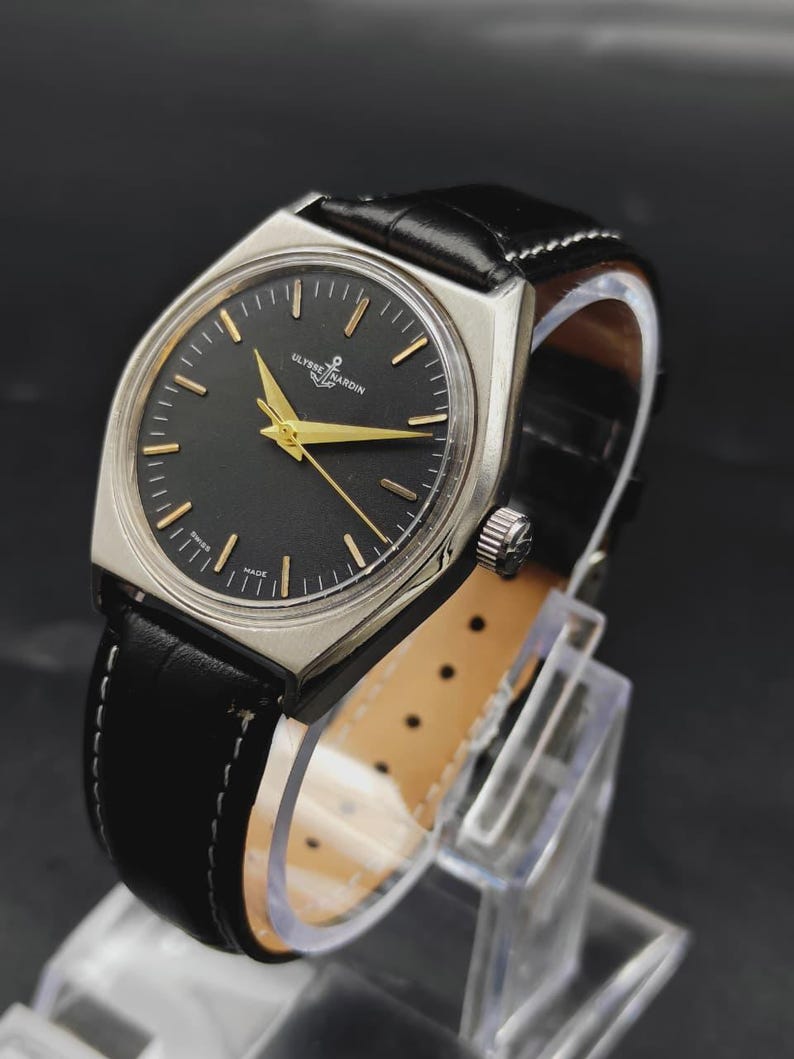 May include: A Ulysse Nardin wristwatch with a black dial, gold-tone hands, and gold-tone hour markers. The watch features a silver-tone metal case and a black leather strap with white stitching. The text Ulysse Nardin is printed on the dial.