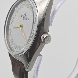 May include: A silver-toned Jaeger-LeCoultre Club watch with a white face, gold hands, and a brown leather strap. The watch face displays the Jaeger-LeCoultre logo and the word "CLUB". The watch is displayed on a clear stand.