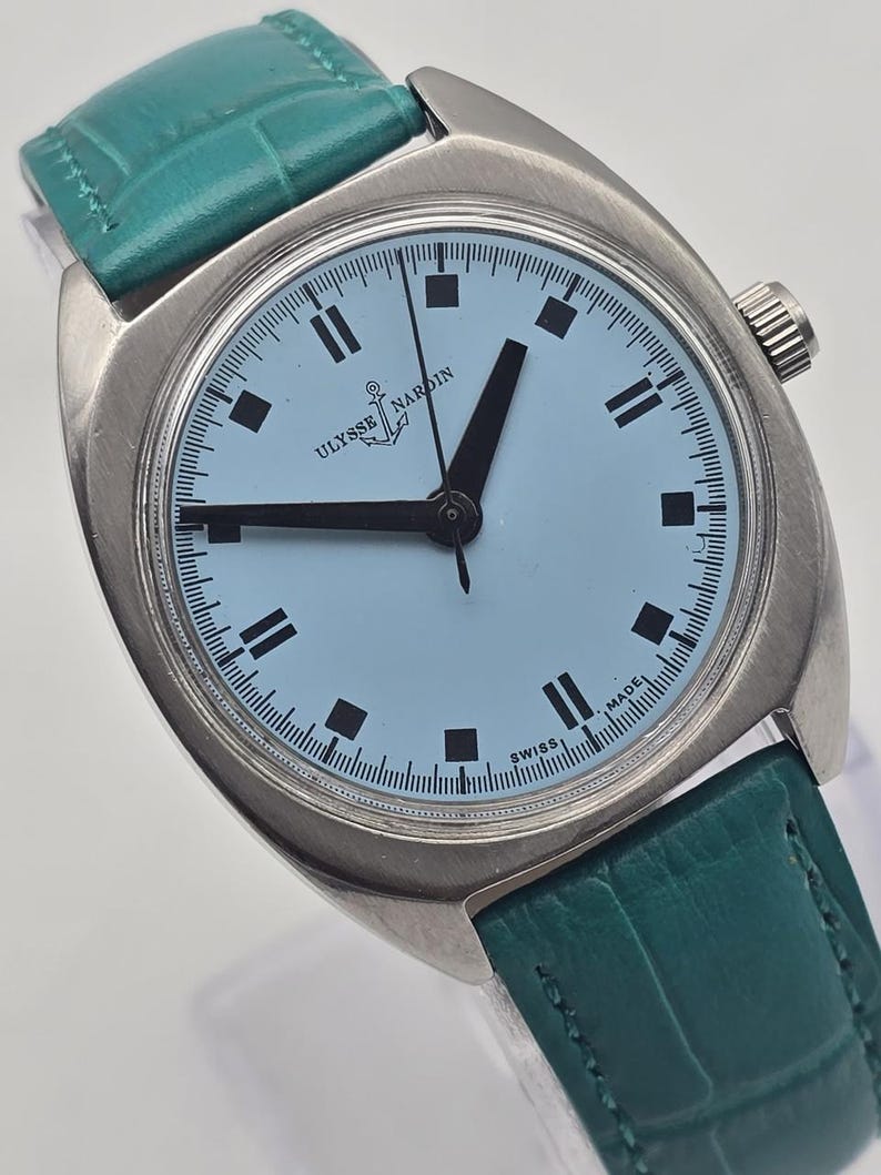 Peut inclure: Une montre-bracelet Ulysse Nardin vintage avec un cadran bleu clair et des aiguilles et marqueurs noirs. La montre a un bo&icirc;tier argent&eacute; et un bracelet en cuir vert d'eau. Le cadran affiche le logo Ulysse Nardin et l'inscription "SWISS MADE".