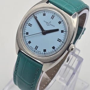 Peut inclure: Une montre Ulysse Nardin vintage avec un cadran bleu clair et des aiguilles noires. La montre a un bo&icirc;tier argent&eacute; et un bracelet en cuir vert d'eau. La montre est pr&eacute;sent&eacute;e sur un support transparent.