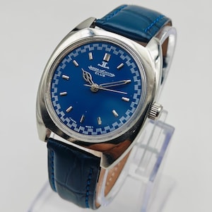 Puede incluir: Un reloj Jaeger-LeCoultre Club plateado con esfera azul y correa de cuero azul. El reloj tiene una esfera redonda con agujas y marcadores plateados. La correa se sujeta con una hebilla plateada. El reloj se exhibe sobre un soporte transparente.
