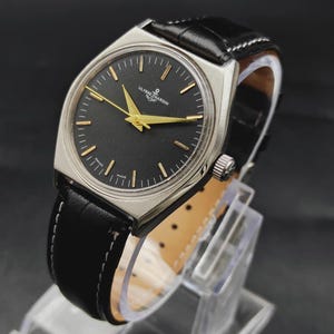 May include: A wristwatch with a silver-toned case, a black dial, and gold-colored hands and hour markers. The dial features the text Ulysse Nardin and an anchor logo. It has a black leather strap with white stitching and a silver crown on the side.