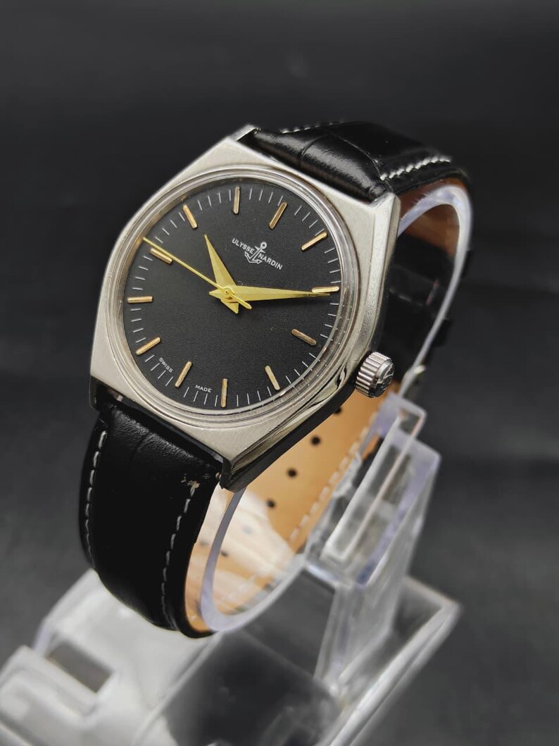 May include: A wristwatch with a silver-toned case, a black dial, and gold-colored hands and hour markers. The dial features the text Ulysse Nardin and an anchor logo. It has a black leather strap with white stitching and a silver crown on the side.