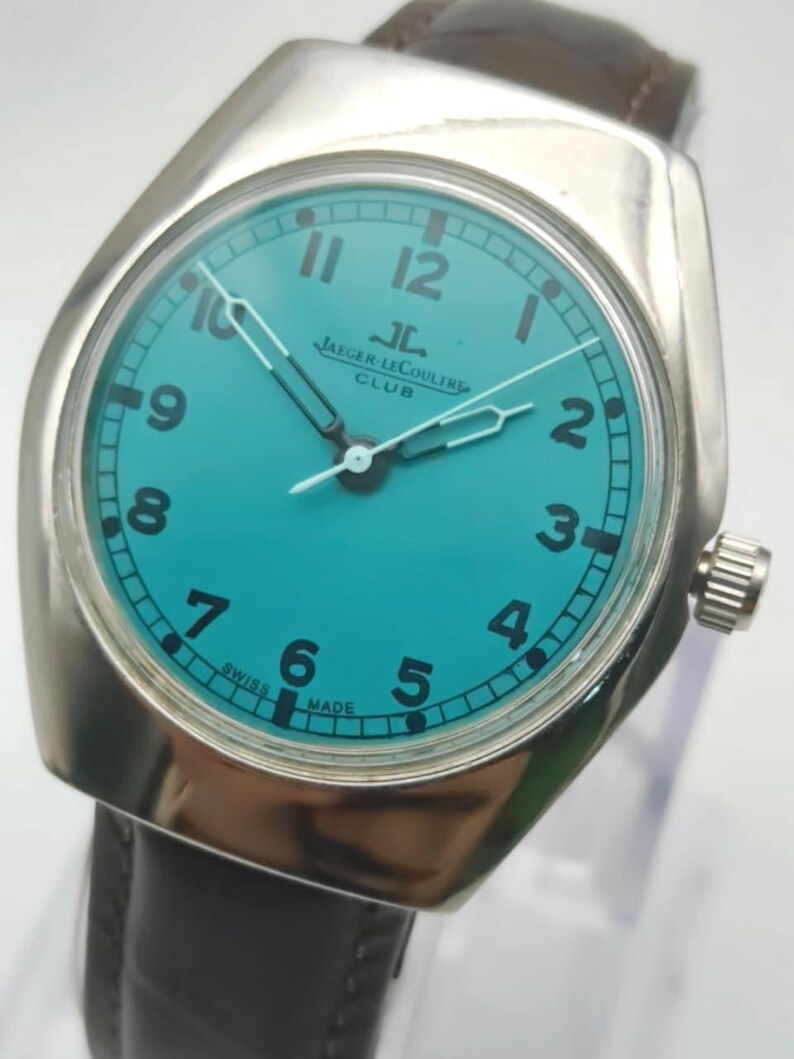 Op de afbeelding: Een vintage Jaeger-LeCoultre Club horloge met een turquoise wijzerplaat en een zilverkleurige kast. Het horloge heeft zwarte cijfers, witte wijzers en een bruine leren band. De tekst "JAEGER-LECOULTRE CLUB" is zichtbaar. Swiss Made.