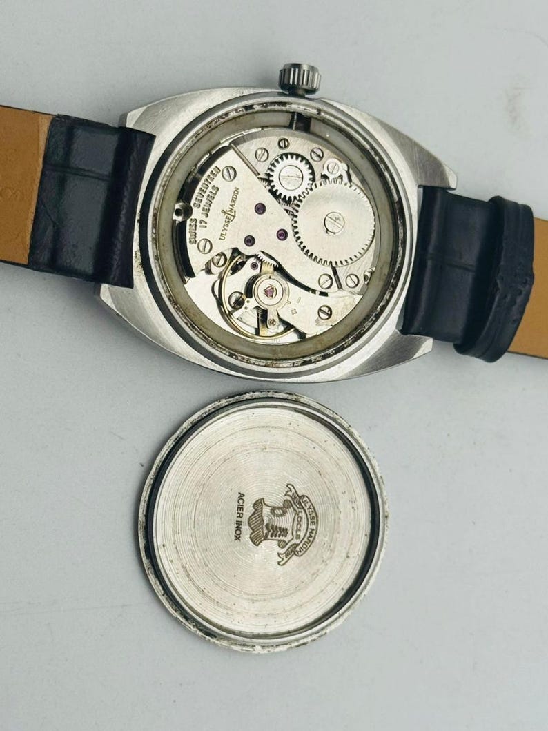 May include: A vintage silver watch with a black leather strap. The watch's back is open, displaying the internal mechanisms, including gears and jewels. The detached back shows the brand's logo and the words "Acier Inox".