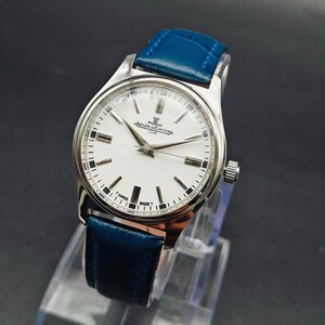 Peut inclure: Une montre-bracelet argent&eacute;e avec un cadran blanc et un bracelet en cuir bleu. Le cadran de la montre comporte des marqueurs et des aiguilles noirs, avec la marque "Jaeger-LeCoultre" visible. La montre est expos&eacute;e sur un support transparent.