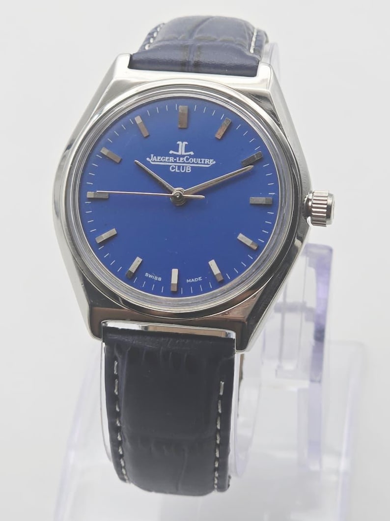May include: A silver-toned Jaeger-LeCoultre Club watch with a blue dial and a black leather strap. The watch face features silver-toned hour markers and hands. The words "JAEGER-LECOULTRE CLUB" are printed on the dial.