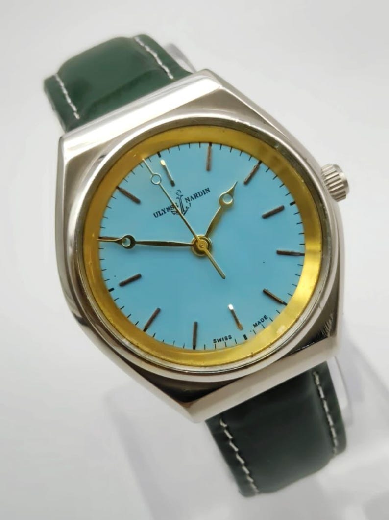 May include: A silver-toned Ulysse Nardin watch with a light blue face and gold-colored accents. The watch has a dark green leather strap. The watch face has black hour markers and hands.