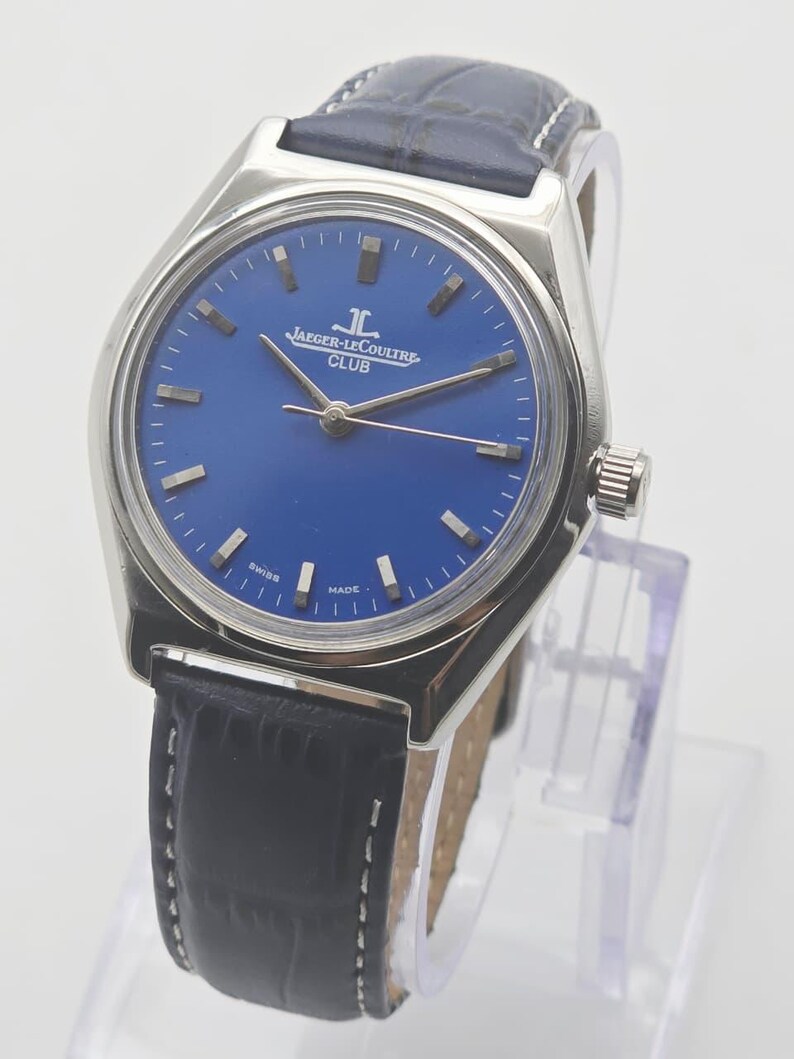 May include: A silver-toned Jaeger-LeCoultre Club watch with a deep blue face and silver hands. The watch has a black leather band with white stitching. The watch face displays the Jaeger-LeCoultre logo and the word "CLUB".