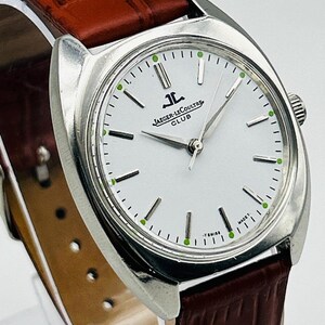 May include: A silver-toned Jaeger-LeCoultre Club watch with a white face, featuring black and green accents. The watch has a brown leather band. The watch face displays the Jaeger-LeCoultre logo and the words "CLUB" and "T SWISS MADE T".
