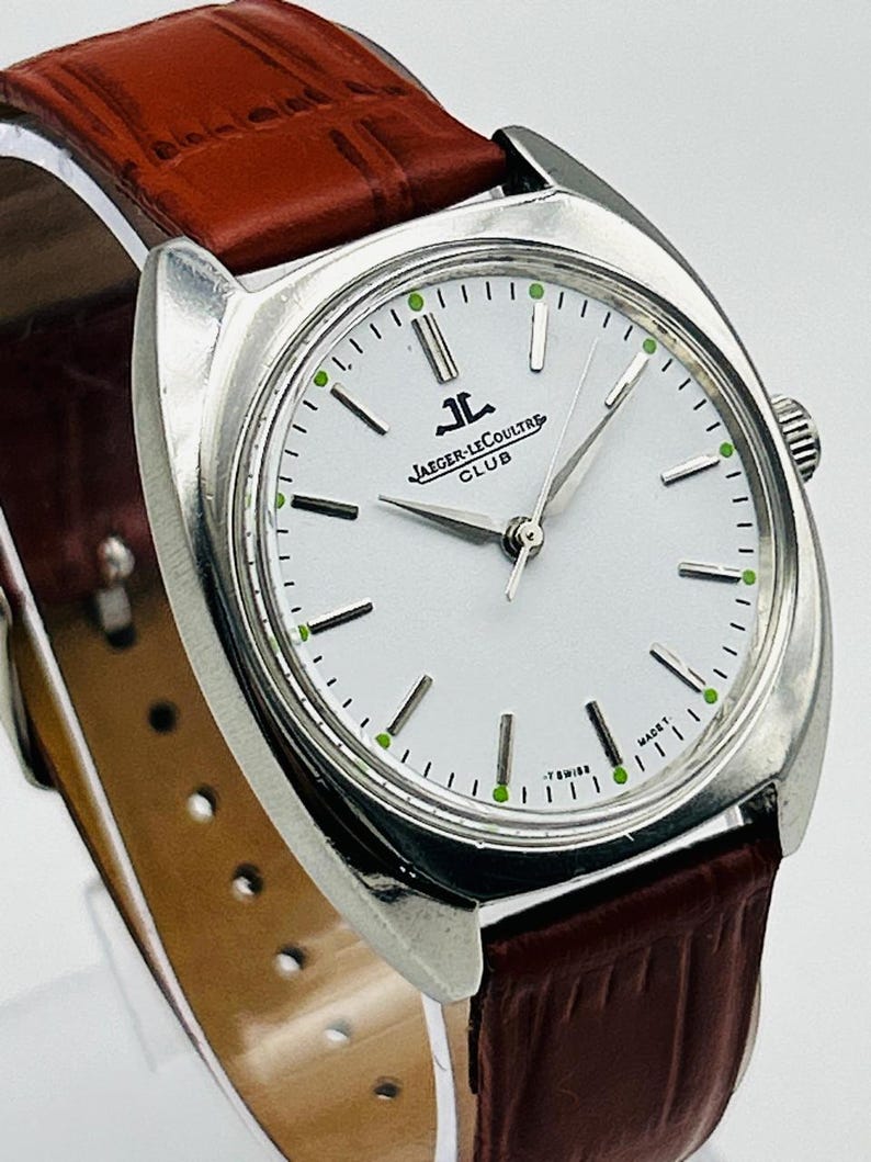 May include: A silver-toned Jaeger-LeCoultre Club watch with a white face, featuring black and green accents. The watch has a brown leather band. The watch face displays the Jaeger-LeCoultre logo and the words "CLUB" and "T SWISS MADE T".