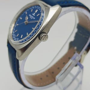 Puede incluir: Un reloj de pulsera plateado con esfera azul y correa de cuero azul. La esfera del reloj tiene agujas e indicadores plateados. El reloj se exhibe sobre un soporte transparente.