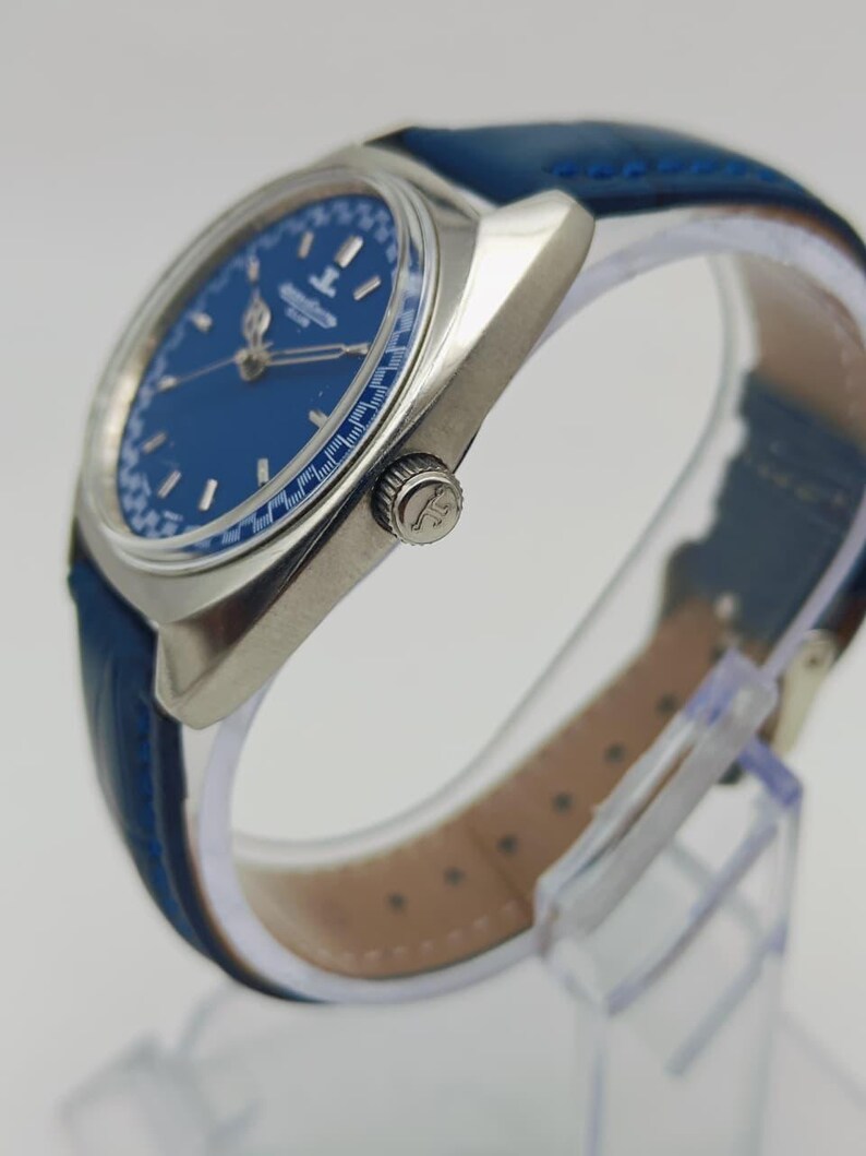 Puede incluir: Un reloj de pulsera plateado con esfera azul y correa de cuero azul. La esfera del reloj tiene agujas e indicadores plateados. El reloj se exhibe sobre un soporte transparente.